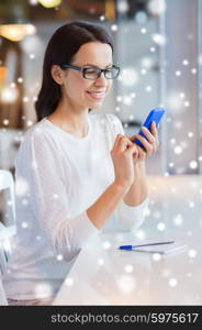 business, people, technology and lifestyle concept - smiling young woman in eyeglasses texting message with smartphone at cafe over snow effect