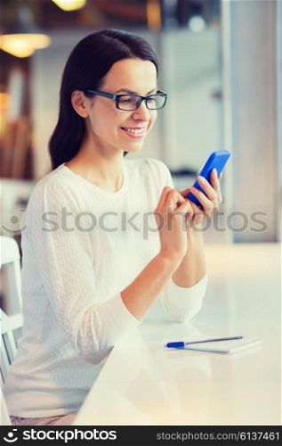 business, people, technology and lifestyle concept - smiling young woman in eyeglasses texting message with smartphone at cafe