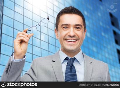 business, people, vision and office concept - happy smiling businessman in eyeglasses and suit over office building background