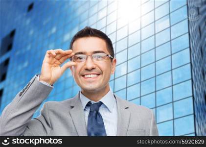 business, people, vision and office concept - happy smiling businessman in eyeglasses and suit over office building background