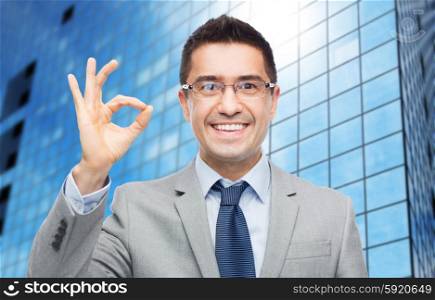 business, people, vision and success concept - happy smiling businessman in eyeglasses and suit showing ok sign over office building background
