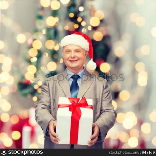 business, presents and people concept - smiling senior man in suit and santa helper hat with gift over christmas tree lights background