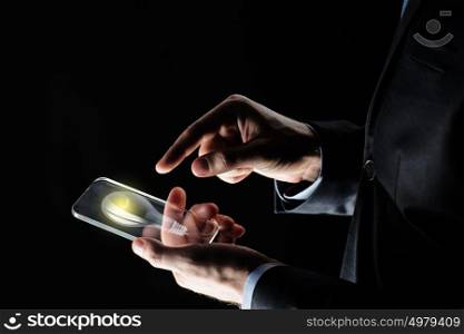 business, solution, idea, people and future technology concept - close up of businessman hands with lightbulb on transparent smartphone screen over black background. businessman with lightbulb on smartphone