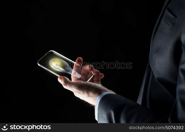 business, solution, idea, people and future technology concept - close up of businessman hand with lightbulb on transparent smartphone screen over black background. businessman with lightbulb on smartphone