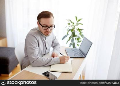 business, startup and people concept - businessman or creative male worker writing to notebook or diary and looking at smartphone at home office