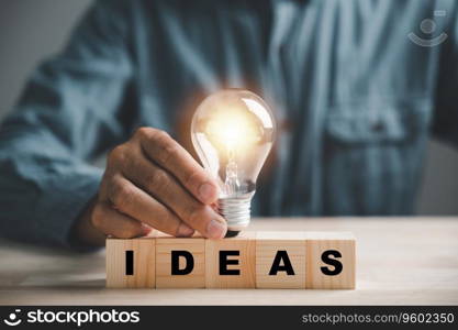 Business startup concept, A mans hand holds a light bulb while word Ideas is displayed on a wooden block. Illustrates power of innovation, inspiration and creative thinking in business development.