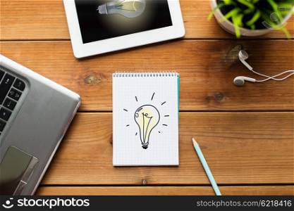 business, startup, idea and technology concept - close up of notebook with light bulb, laptop and tablet pc computer with