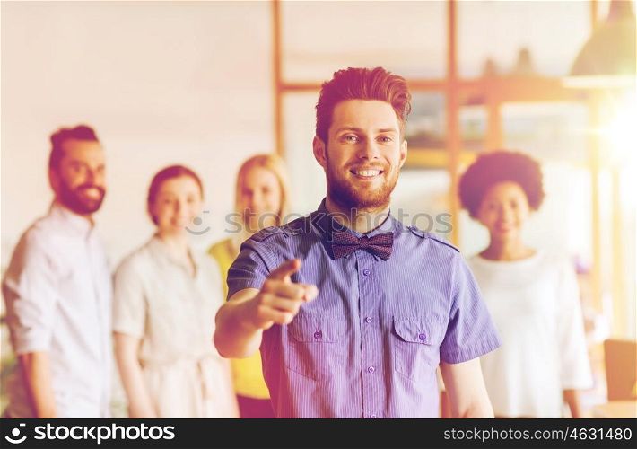 business, startup, people, gesture and teamwork concept - happy young man with beard and bow tie pointing finger at you over creative team in office