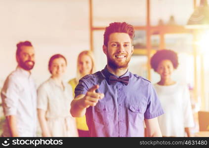 business, startup, people, gesture and teamwork concept - happy young man with beard and bow tie pointing finger at you over creative team in office