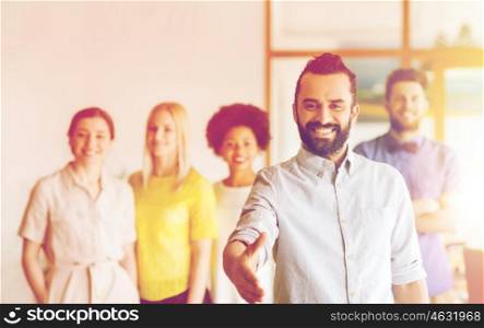 business, startup, people, gesture and teamwork concept - happy young man with beard greeting by handshake over creative team in office