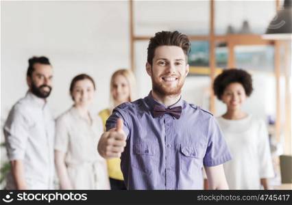 business, startup, people, gesture and teamwork concept - happy young man with beard and bow tie showing thumbs up over creative team in office