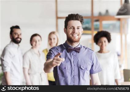 business, startup, people, gesture and teamwork concept - happy young man with beard and bow tie pointing finger at you over creative team in office