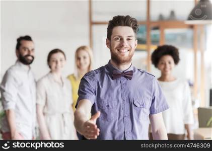business, startup, people, gesture and teamwork concept - happy young man with beard and bow tie greeting by handshake over creative team in office