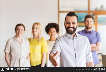 business, startup, people, gesture and teamwork concept - happy young man with beard greeting by handshake over creative team in office
