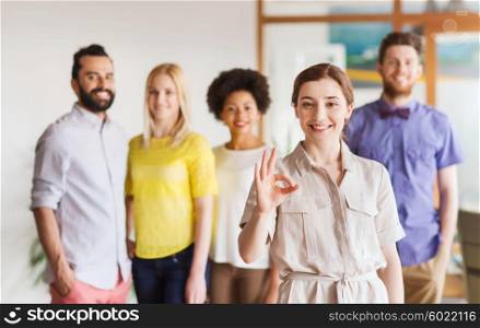business, startup, people, gesture and teamwork concept - happy young woman showing ok hand sig over creative team in office
