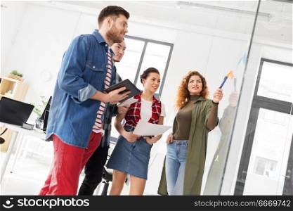 business, startup, planning and people concept - happy creative team discussing something on glass board at office. creative team looking at glass board at office. creative team looking at glass board at office