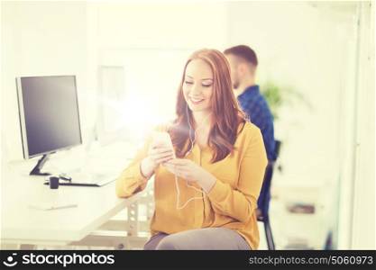 business, startup, technology and people concept - happy businesswoman or creative worker with earphones listening to music on smartphone at office. woman with earphones and smartphone at office