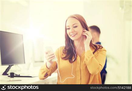 business, startup, technology and people concept - happy businesswoman or creative worker with earphones listening to music on smartphone at office. woman with earphones and smartphone at office