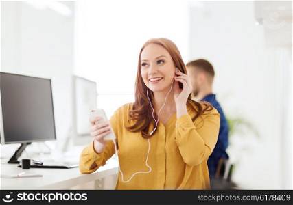 business, startup, technology and people concept - happy businesswoman or creative worker with earphones listening to music on smartphone at office
