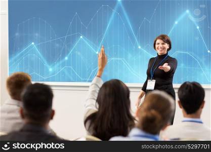 business, statistics and people concept - smiling businesswoman or lecturer with diagram chart on projection screen answering questions at conference presentation or lecture. group of people at business conference or lecture