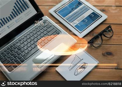 business, statistics and technology concept - close up of on laptop computer, tablet pc, notebook and eyeglasses with charts on wooden table