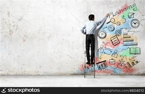 Business strategy and planning . Back view of businessman standing on ladder and drawing sketches on wall
