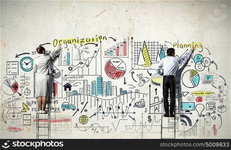 Business strategy seminar. Back view of businessman and businesswoman standing on ladder and drawing sketches on wall