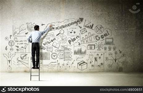Business strategy seminar. Back view of businessman standing on ladder and drawing sketches on wall