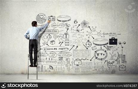 Business strategy seminar. Back view of businessman standing on ladder and drawing sketches on wall