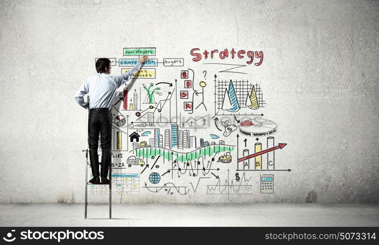 Business strategy seminar. Back view of businessman standing on ladder and drawing sketches on wall