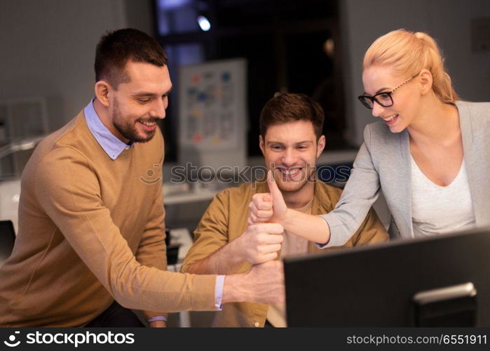 business, success and teamwork concept - happy coworkers with computer working late at night office making thumbs up gesture. business team making thumbs up gesture at office. business team making thumbs up gesture at office