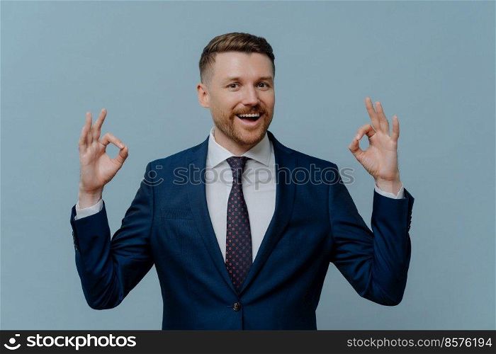Business success. Happy positive businessman or satisfied male entrepreneur in suit showing ok sign or okay gesture and smiling at camera while standing against light steel blue background. Happy businessman showing ok sign and smiling