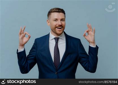 Business success. Happy positive businessman or satisfied male entrepreneur in suit showing ok sign or okay gesture and smiling at camera while standing against light steel blue background. Happy businessman showing ok sign and smiling