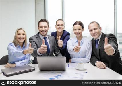 business, success, technology and office concept - smiling business team with laptop computer, papers and coffee showing thumbs up in office