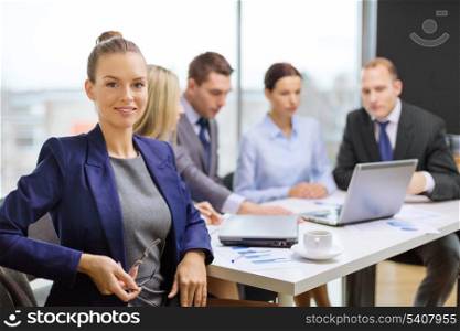 business, technology and office concept - smiling businesswoman with eyeglasses in office with team on the back