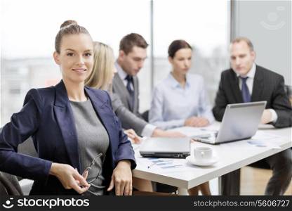 business, technology and office concept - smiling businesswoman with eyeglasses in office with team on the back
