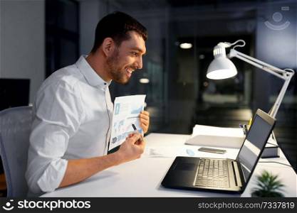 business, technology and people concept - businessman with charts having video chat on laptop computer at night office. businessman having video chat at night office