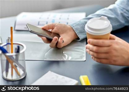 business, technology and people concept - close up of hands using smartphone and drinking takeaway coffee from paper cup at office. woman with coffee using smartphone at office