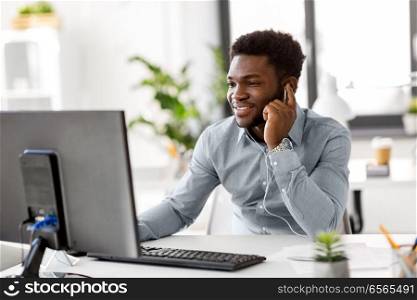 business, technology and people concept - happy african american businessman with earphones and computer computer listening to music at office. businessman with earphones and computer at office