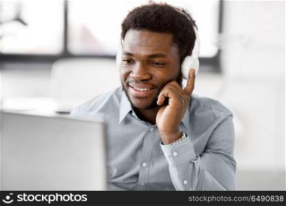business, technology and people concept - happy african american businessman with headphones and computer listening to music at office. businessman with headphones and computer at office. businessman with headphones and computer at office