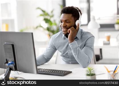 business, technology and people concept - happy african american businessman with headphones and computer computer listening to music at office. businessman with headphones and computer at office. businessman with headphones and computer at office