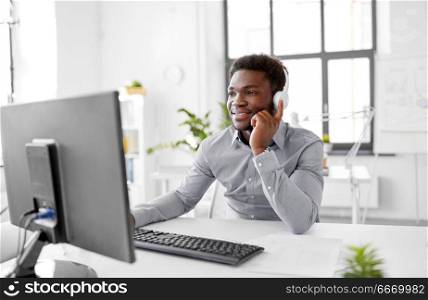 business, technology and people concept - happy african american businessman with headphones and computer listening to music at office. businessman with headphones and computer at office. businessman with headphones and computer at office