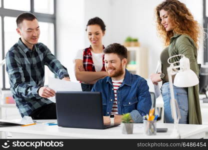 business, technology and people concept - happy creative team with laptop computer and papers working at office. creative team with laptop working at office. creative team with laptop working at office