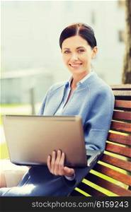 business, technology and people concept - young smiling woman with laptop computer sitting on bench in city