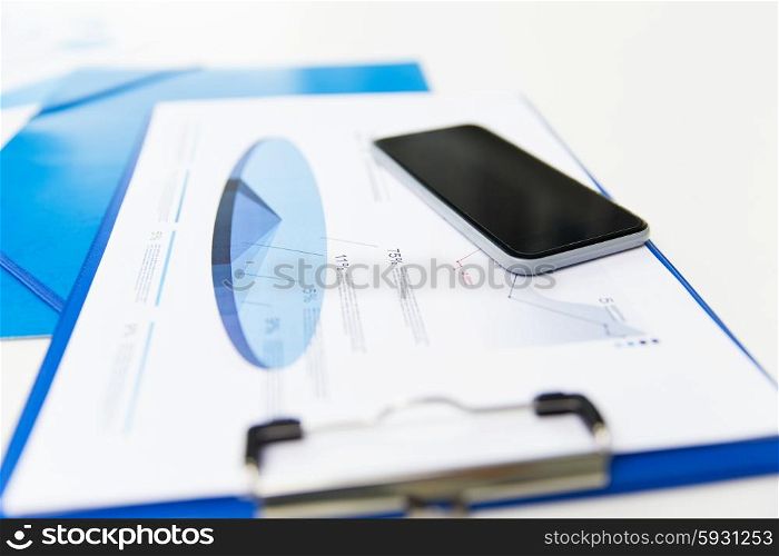 business, technology and statistics concept - close up of smartphone and clipboard with charts on office table