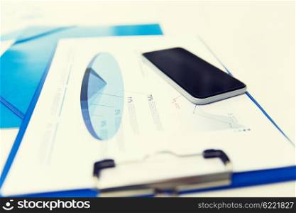 business, technology and statistics concept - close up of smartphone and clipboard with charts on office table