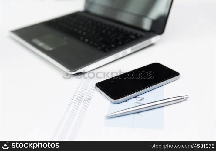 business, technology and statistics concept - close up of smartphone, laptop computer and chart with pen on office table