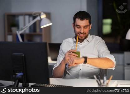 business, technology and time management concept - happy smiling businessman drinking green smoothie and looking at smart watch at nigh office. happy businessman with smart watch at nigh office