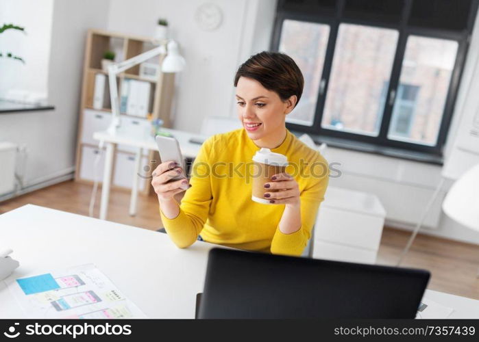 business, technology and user interface design concept - smiling businesswoman or ui designer using smartphone and drinking takeaway coffee from paper cup at office. happy woman with coffee using smartphone at office