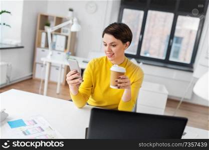 business, technology and user interface design concept - smiling businesswoman or ui designer using smartphone and drinking takeaway coffee from paper cup at office. happy woman with coffee using smartphone at office